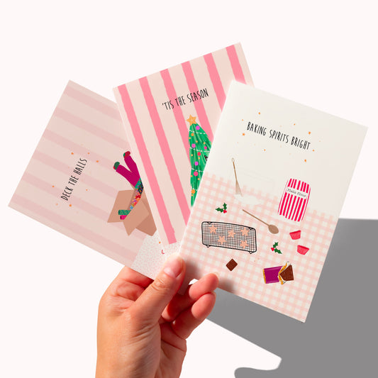 Three illustrated greeting cards are fanned out in a hand against a clean white background. The cards feature pastel pink and white designs with baking-themed illustrations, including a striped pattern, a green Christmas tree, and small holiday accents.