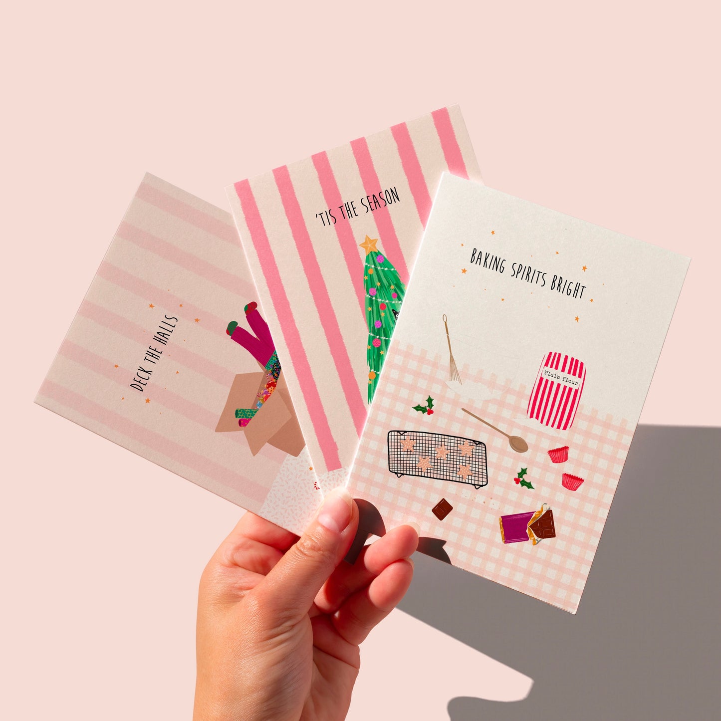 Three girly Christmas-themed greeting cards held by a hand against a pink background