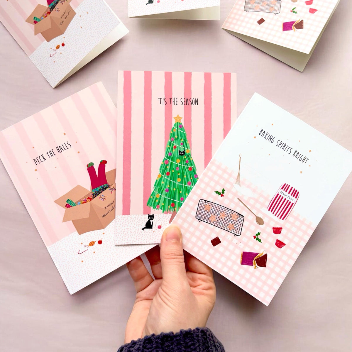 A hand holds three girly Christmas-themed greeting cards against a soft pink background. The cards feature cute holiday illustrations, including a green Christmas tree with black cats playing, a girl’s legs sticking up out of a box of decorations, and festive baking, with playful text like 'T’is the season' and 'Baking spirits bright!'.