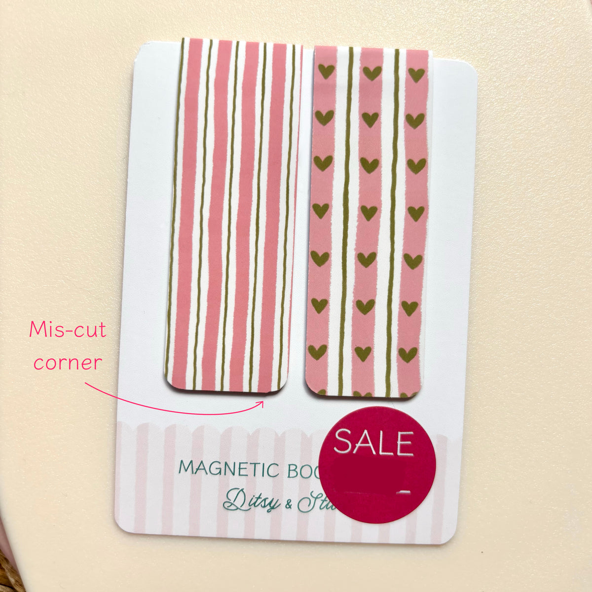 A white card holding two decorative rectangular magnetic pieces with pink and olive-green striping and heart motifs, photographed on a beige surface. Pink text reads 'Mis-cut corner,' and a magenta 'SALE' sticker is overlaid near the bottom.