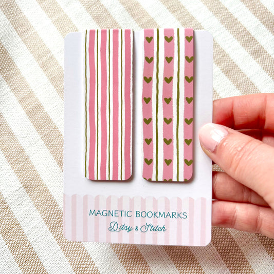 Two magnetic bookmarks with striped and heart patterns in pink and green on a card held by a hand against a striped background.
