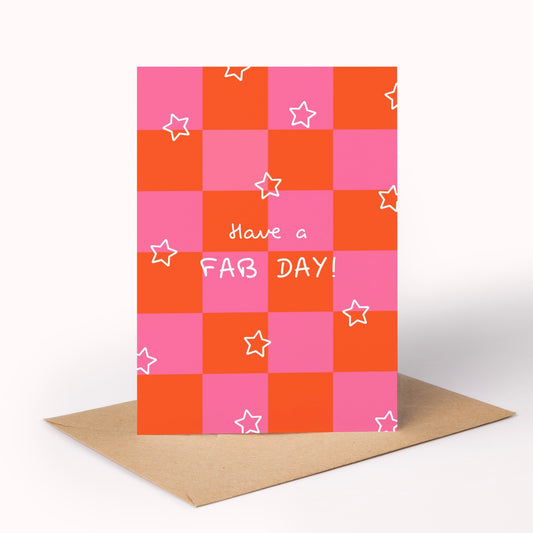 A greeting card with a pink and orange checkerboard pattern, decorated with small white outlined stars and the text 'Have a FAB DAY!' in white script and block letters. The card is standing upright on a brown kraft envelope against a clean white background.