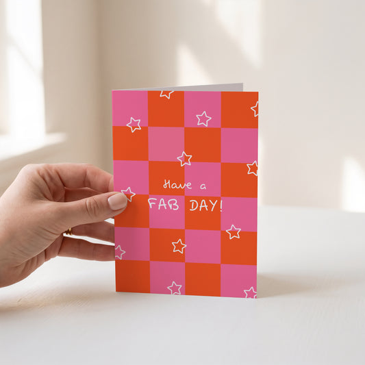 A hand holds a greeting card with a pink and orange checkerboard pattern, white star outlines, and the text 'Have a FAB DAY!' against a softly lit indoor background.