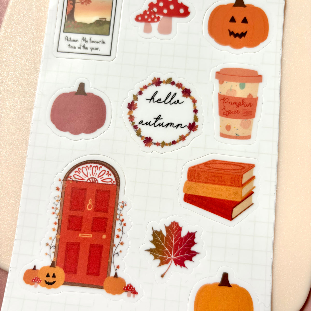 A sheet of autumn-themed stickers on a white grid background, featuring pumpkins, a red door, a maple leaf, books, a coffee cup, mushrooms, and a 'hello autumn' wreath.