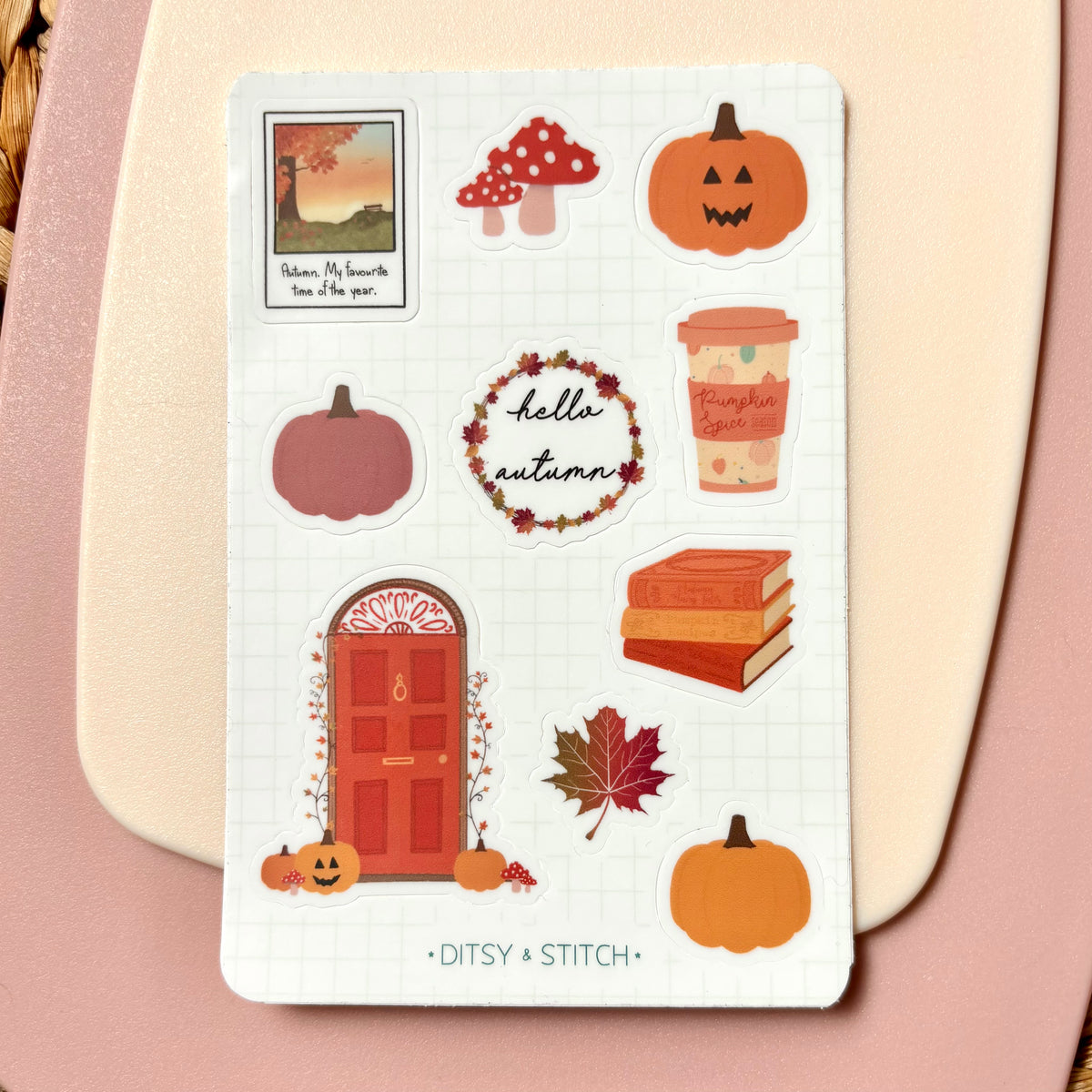 A sheet of autumn-themed planner stickers on a white grid background, placed on a beige card over a pink surface. The stickers include pumpkins, mushrooms, a coffee cup, books, a door, leaves, and a 'hello autumn' wreath.