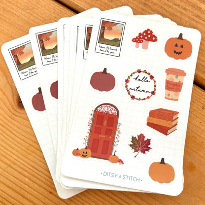 A stack of white sticker sheets featuring autumn-themed illustrations, including pumpkins, a red door, mushrooms, a coffee cup, books, a leaf, and a 'hello autumn' wreath, placed on a wooden surface. The brand name 'Ditsy + Stitch' is printed at the bottom of the sheets.
