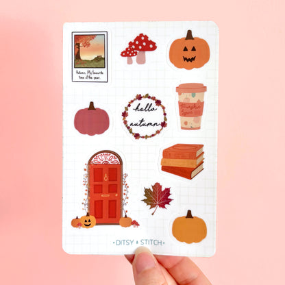A hand holds a white sticker sheet featuring autumn-themed illustrations including pumpkins, a red door, mushrooms, a coffee cup, a leaf, and a wreath with the text 'hello autumn'. The brand name 'DITSY STITCH' is printed at the bottom of the sheet, set against a solid light pink background.