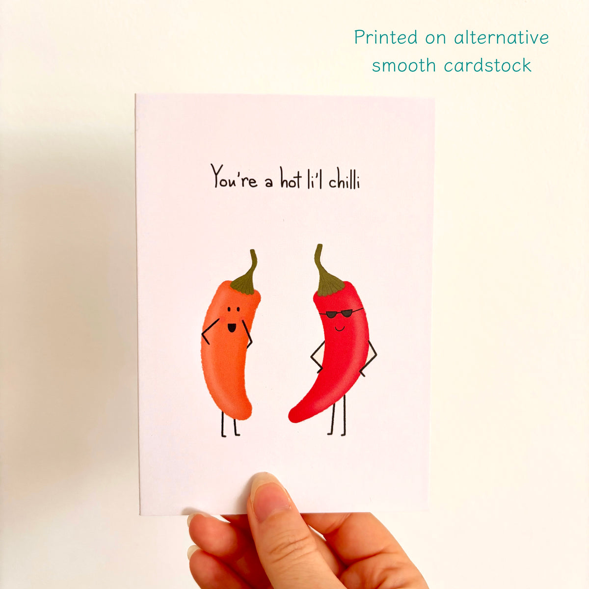 A hand holds a square greeting card with a humorous chilli pepper illustration. The card reads 'You're a hot lil' chilli' with one orange chilli and one red chilli character.