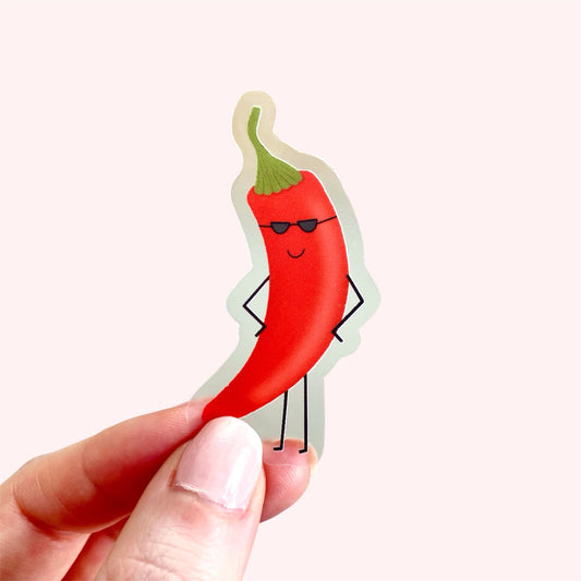 A hand holds a die-cut sticker of a red chilli pepper character wearing black sunglasses, against a solid light pink background.