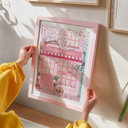 Person holding a framed illustration of a pink patisserie with cherry blossoms.
