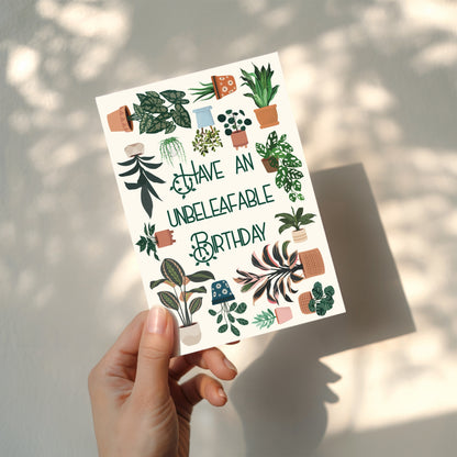 Hand holding a card with plant illustrations and text 'Have an unbeleafable birthday' against a neutral background.
