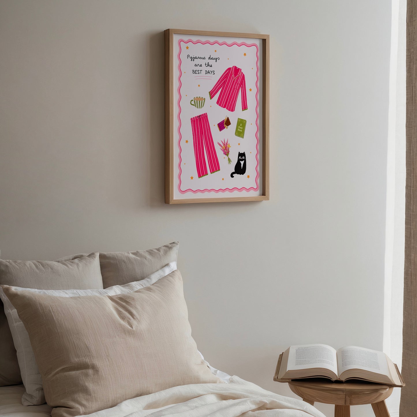Framed pink pyjamas artwork on a wall above a bed with pillows and an open book on a small table.