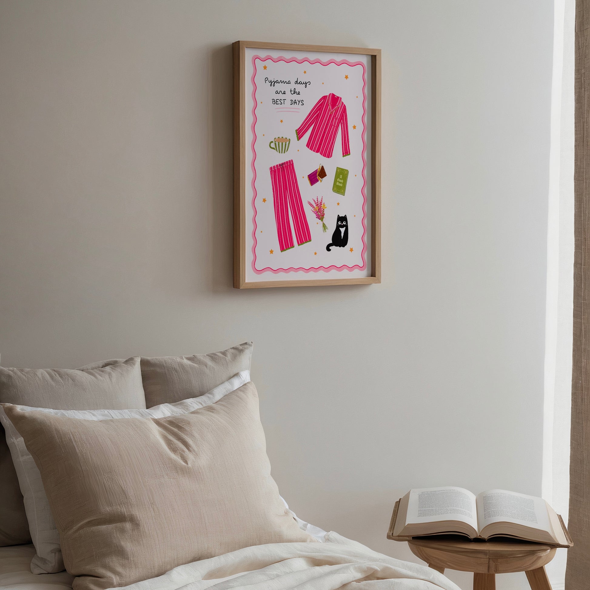 Framed pink pyjamas artwork on a wall above a bed with pillows and an open book on a small table.