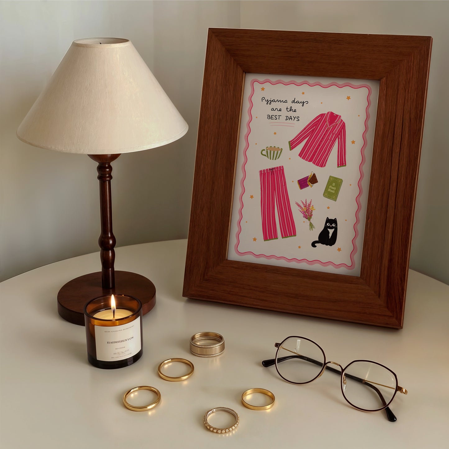 Framed artwork with pink pyjamas design, candle, glasses, and rings on a surface.