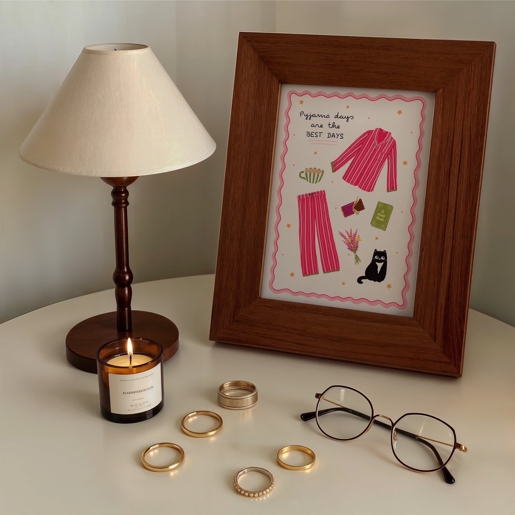 Framed artwork with pink pyjamas design, candle, glasses, and rings on a surface.