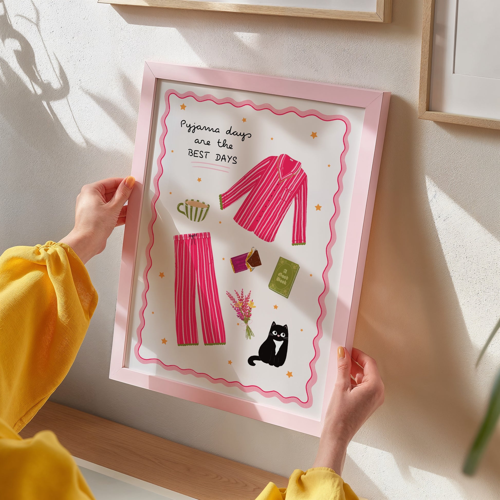 Person holding a framed illustration of pyjama-themed artwork with pink pyjamas and cat.