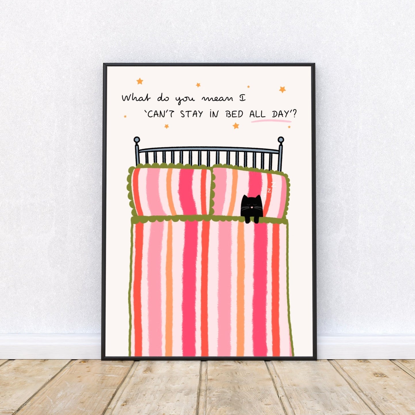 Framed artwork of a bed with striped bedding and a cat, on a white wall, with a quote 'What do you mean I 'can't stay in bed all day'' surrounded by stars.