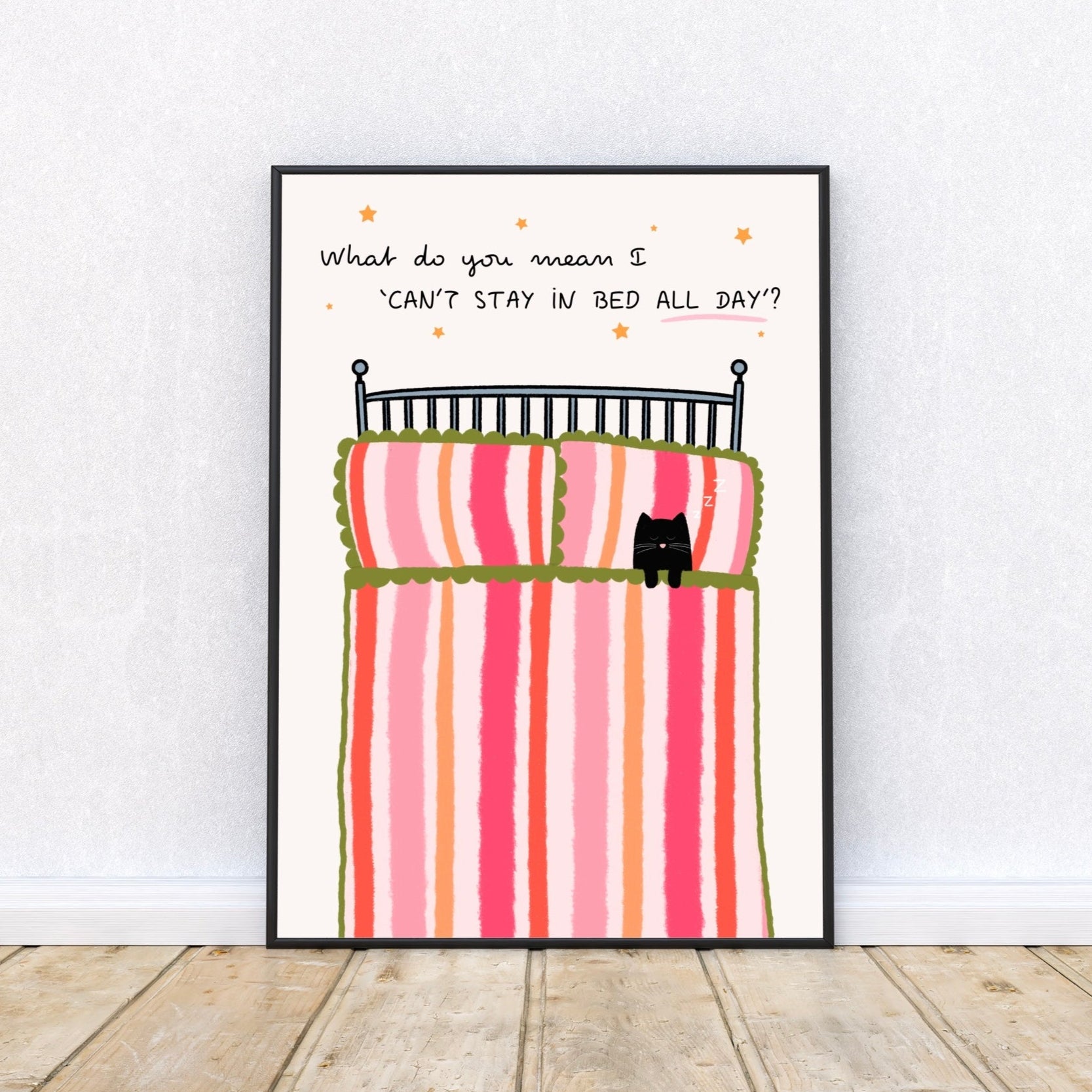 Framed artwork of a bed with striped bedding and a cat, on a white wall, with a quote 'What do you mean I 'can't stay in bed all day'' surrounded by stars.