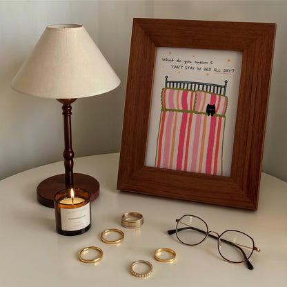 Framed artwork with a bed illustration,  a lamp, candle, glasses, and gold rings on a bedside table.