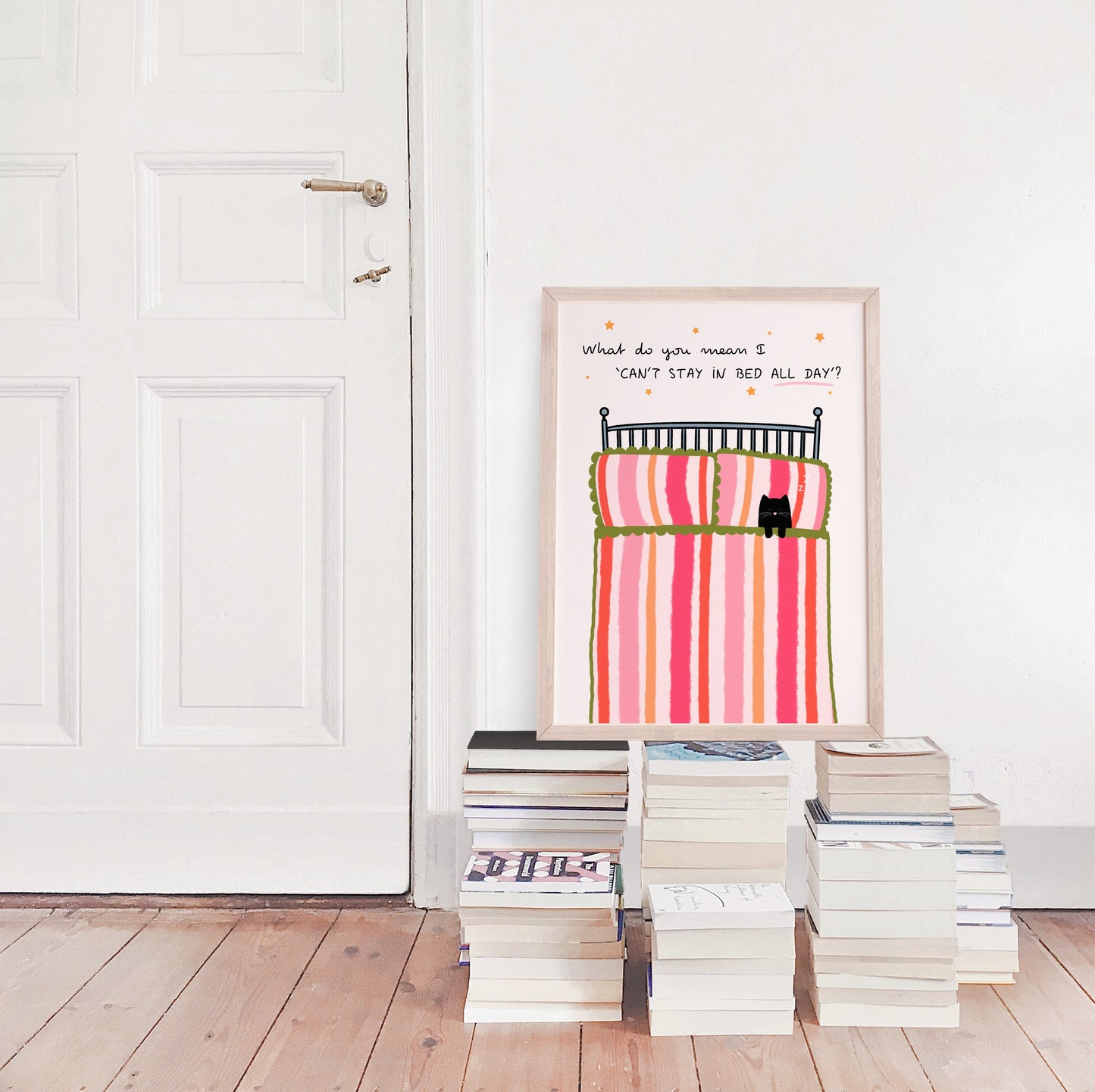 Framed cat in pink bed artwork on a wall with books stacked below