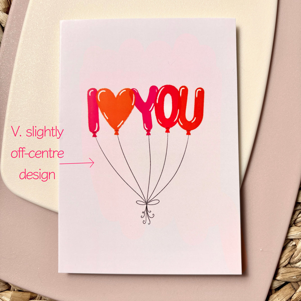 A square greeting card with a white background featuring the text 'I <heart> YOU' in red, pink and orange balloon shapes, resting on a beige background. Pink handwritten text on the left notes that the crd design is v. slightly off centre.