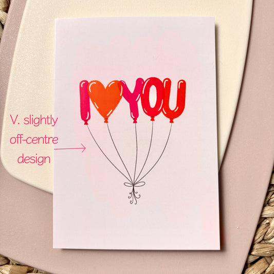 A square greeting card with a white background featuring the text 'I <heart> YOU' in red, pink and orange balloon shapes, resting on a beige background. Pink handwritten text on the left notes that the crd design is v. slightly off centre.