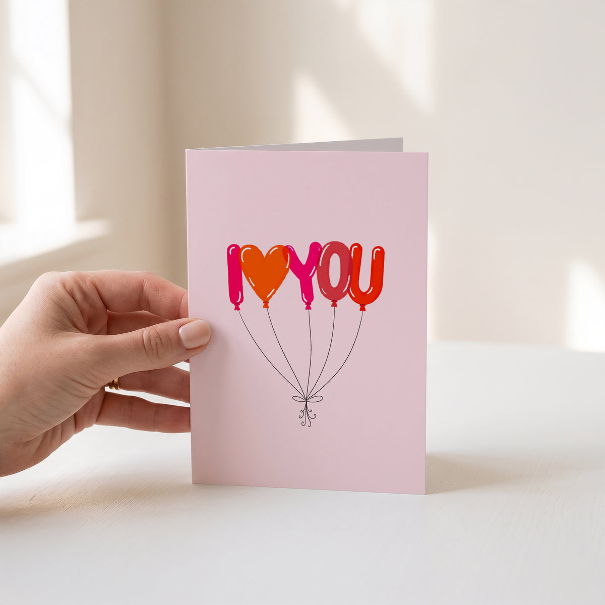 A hand holds a light pink greeting card featuring the phrase 'I <heart> YOU' in balloon-style letters in pink, orange, and red, with thin black strings below. The background is a softly lit, warm off-white interior.