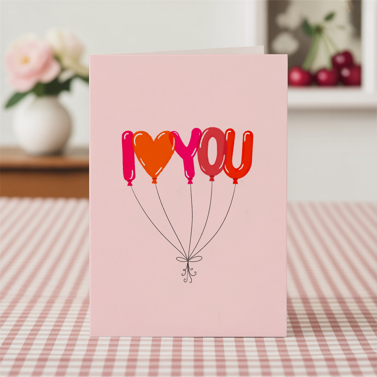 A pink greeting card standing upright on a red-and-white checkered tablecloth, featuring the phrase 'I <heart> YOU' in balloon-style letters in pink, orange, and red, with thin black strings below.