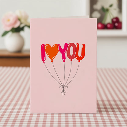 A pink greeting card standing upright on a red-and-white checkered tablecloth, featuring the phrase 'I <heart> YOU' in balloon-style letters in pink, orange, and red, with thin black strings below.