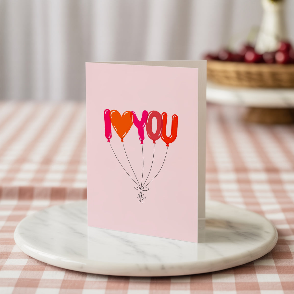 A pink greeting card with the phrase 'I <heart> YOU' in balloon-style letters, standing on a round white marble tray over a pink-and-white checkered tablecloth.