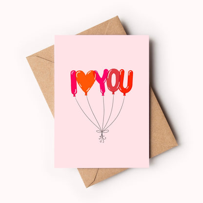A pink greeting card with a balloon-style 'I <heart> YOU' design, shown with a brown kraft envelope on a clean white background.