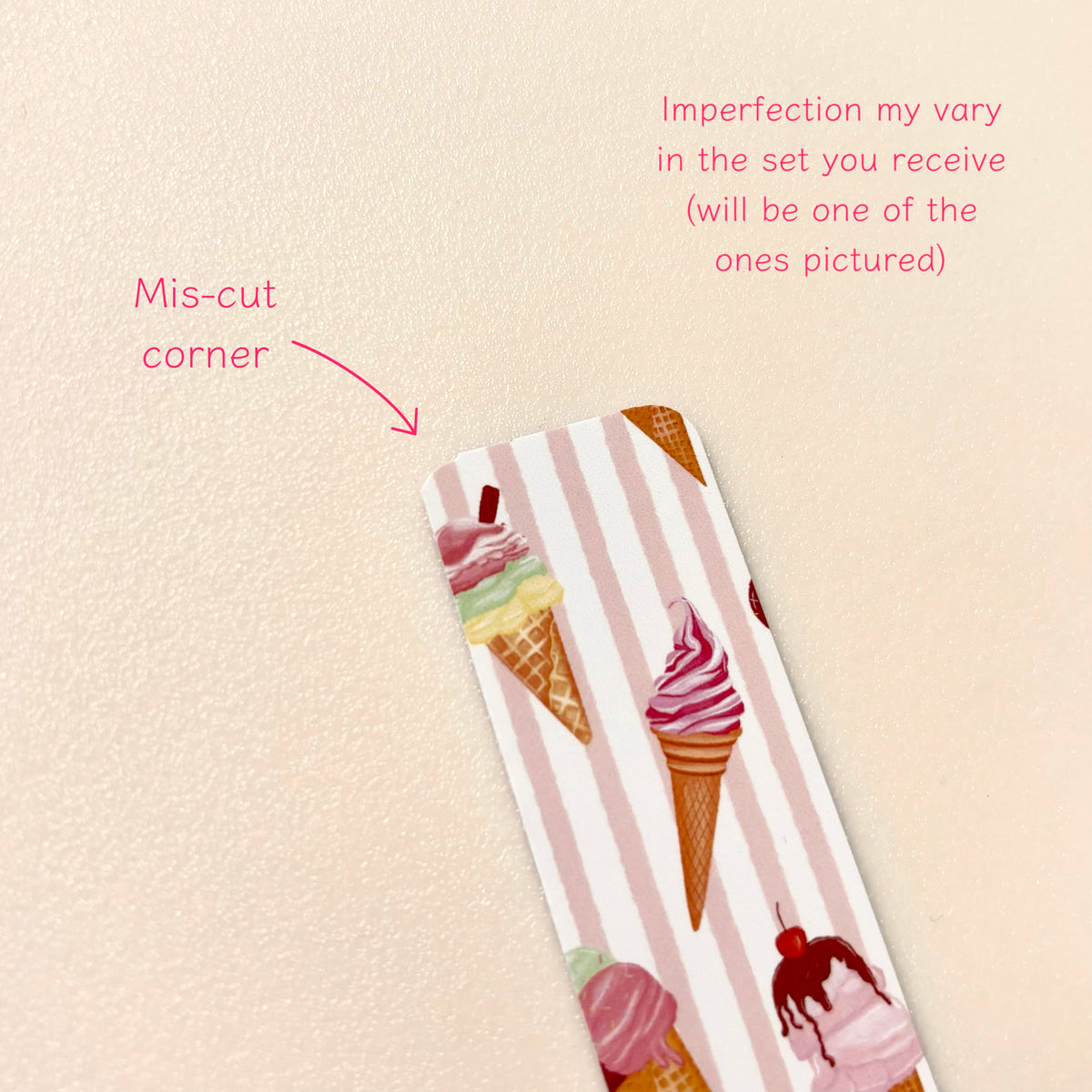 A close-up of a magnetic bookmark with pink and white vertical stripes and colourful ice cream cone illustrations, placed on a light beige textured background. Pink text and arrow call out a mis-cut corner and note variation in the set.