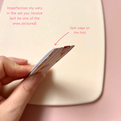 A hand holds a magnetic bookmark showing a split edge, with pink text callouts explaining that imperfections vary by set and pointing to the split edge on the fold. The background is a soft pink surface.