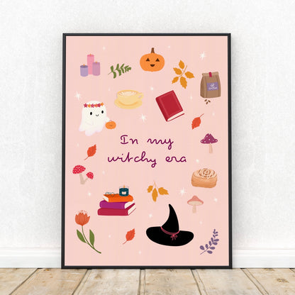 A framed art print featuring a light pink background with whimsical autumn and witch-themed illustrations, including a pumpkin, ghost, witch hat, books, mushrooms, and the text 'In my witchy era'. The print is displayed leaning against a white wall on a light wood floor.