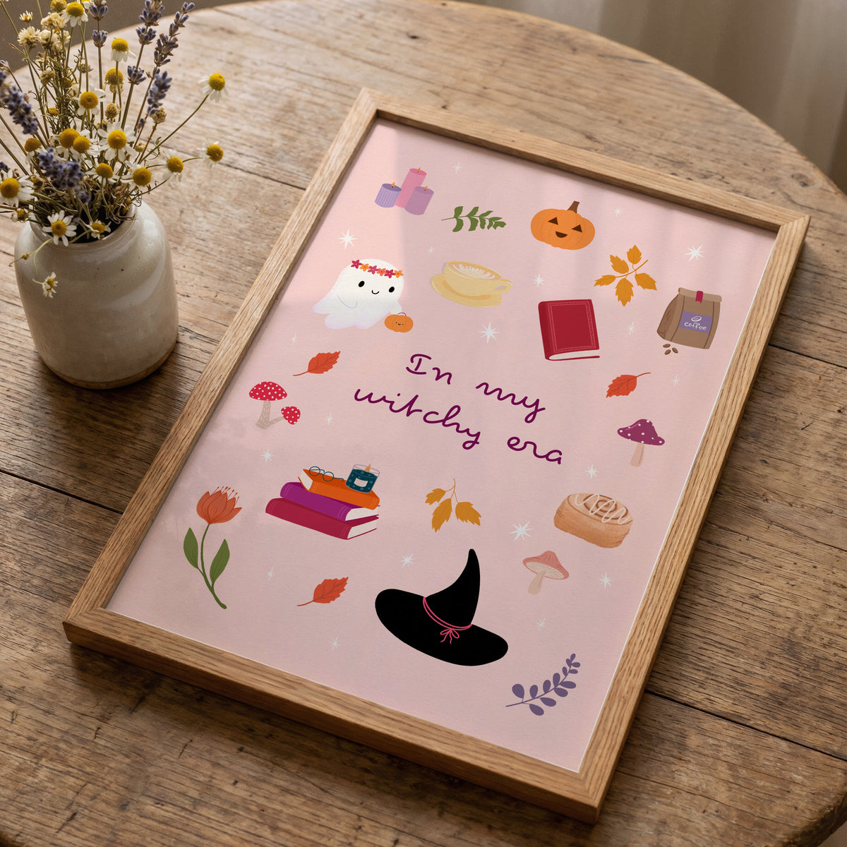 A framed art print with a whimsical witch-themed illustration sits on a rustic wooden table next to a small vase of dried flowers. The print features a light pink background with illustrations of a black witch hat, books, a pumpkin, a ghost, and the text 'In my witchy era'.
