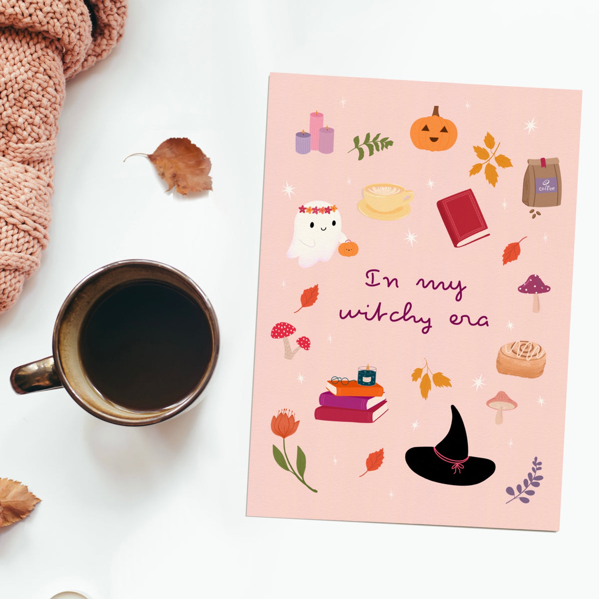 A flat-lay photograph of a pink art print with a witchy autumn theme, styled on a white surface next to a cup of coffee, a knitted scarf, and scattered leaves. The card features illustrations of a ghost, a pumpkin, a witch hat, mushrooms, and books with the text 'In my witchy era'.