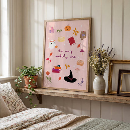 A framed art print featuring a whimsical autumn and witch-themed illustration with the text 'In my witchy era' displayed on a rustic wooden shelf in a cozy bedroom setting.
