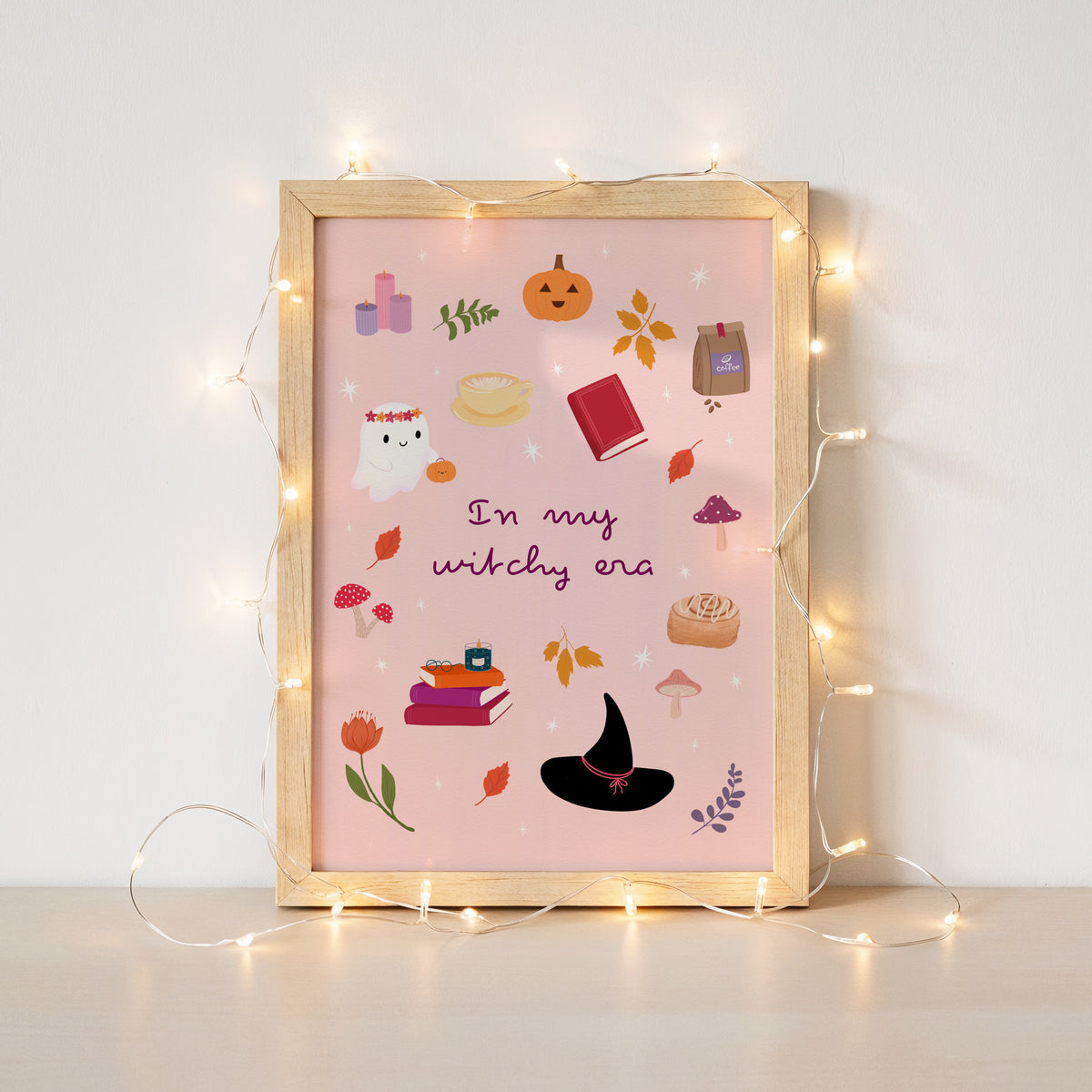 A framed art print with a light wood frame and warm fairy lights draped around it, featuring a pink background with cute Halloween illustrations and the text 'In my witchy era'.