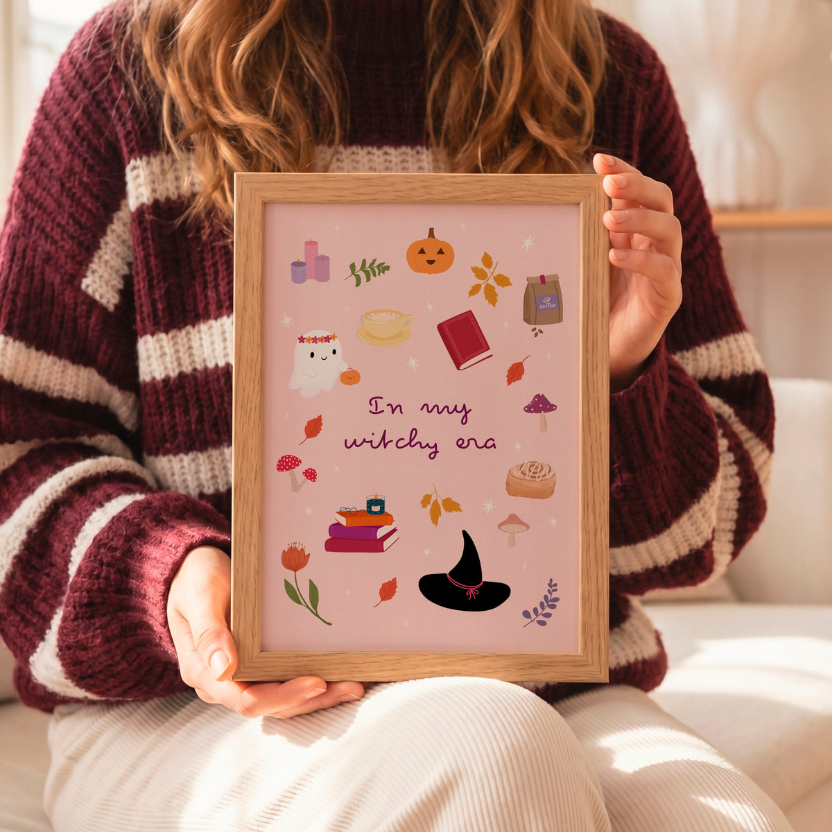 A person in a maroon and cream striped knit sweater holds a framed art print featuring a pink background with cute autumn and witch-themed illustrations and the text 'In my witchy era'.