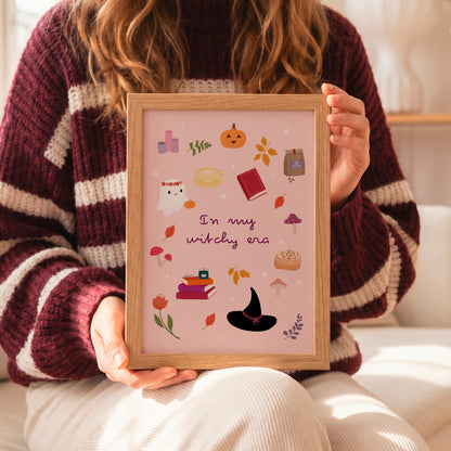 A person in a maroon and cream striped knit sweater holds a framed art print featuring a pink background with cute autumn and witch-themed illustrations and the text 'In my witchy era'.
