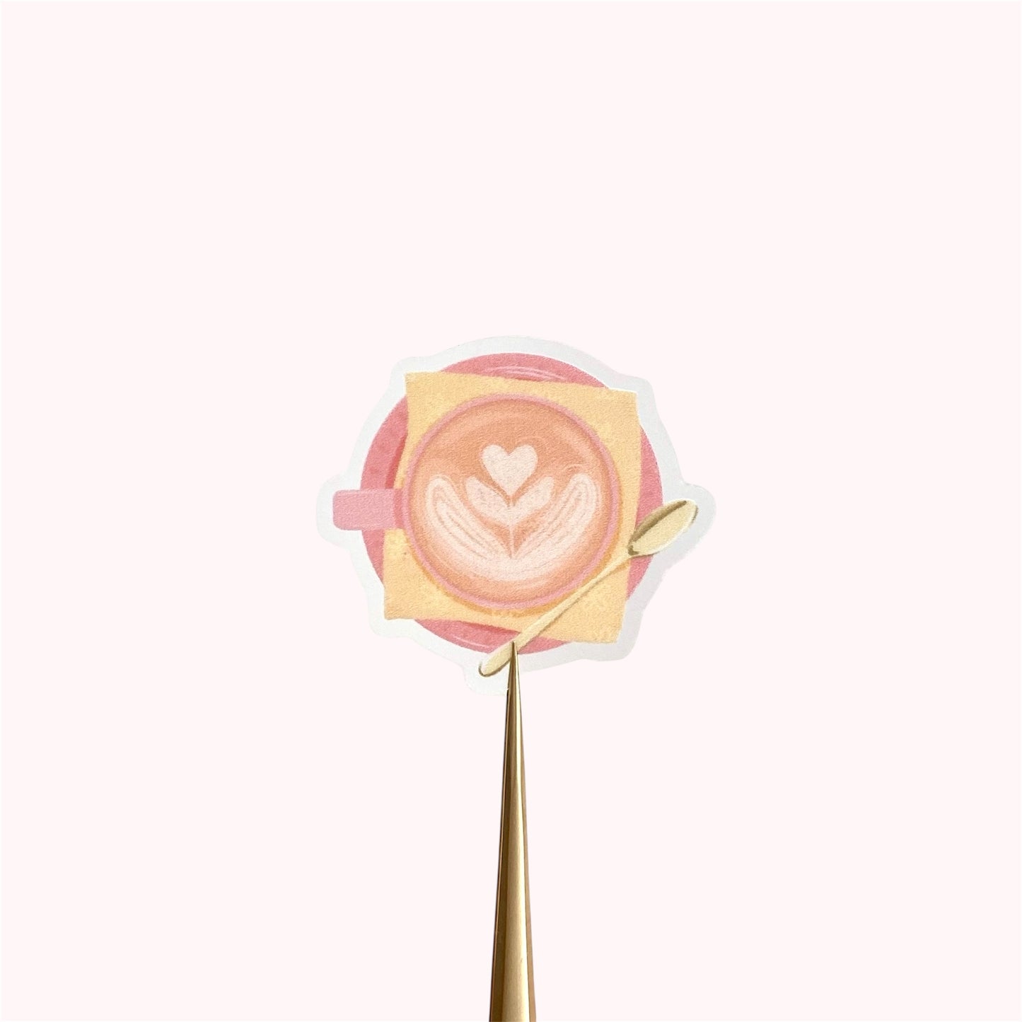 A transparent sticker with a detailed illustration of a latte from above with foamy heart shaped latte art, in a pink mug with a yellow napkin and gold teaspoon.
