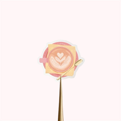 A transparent sticker with a detailed illustration of a latte from above with foamy heart shaped latte art, in a pink mug with a yellow napkin and gold teaspoon.