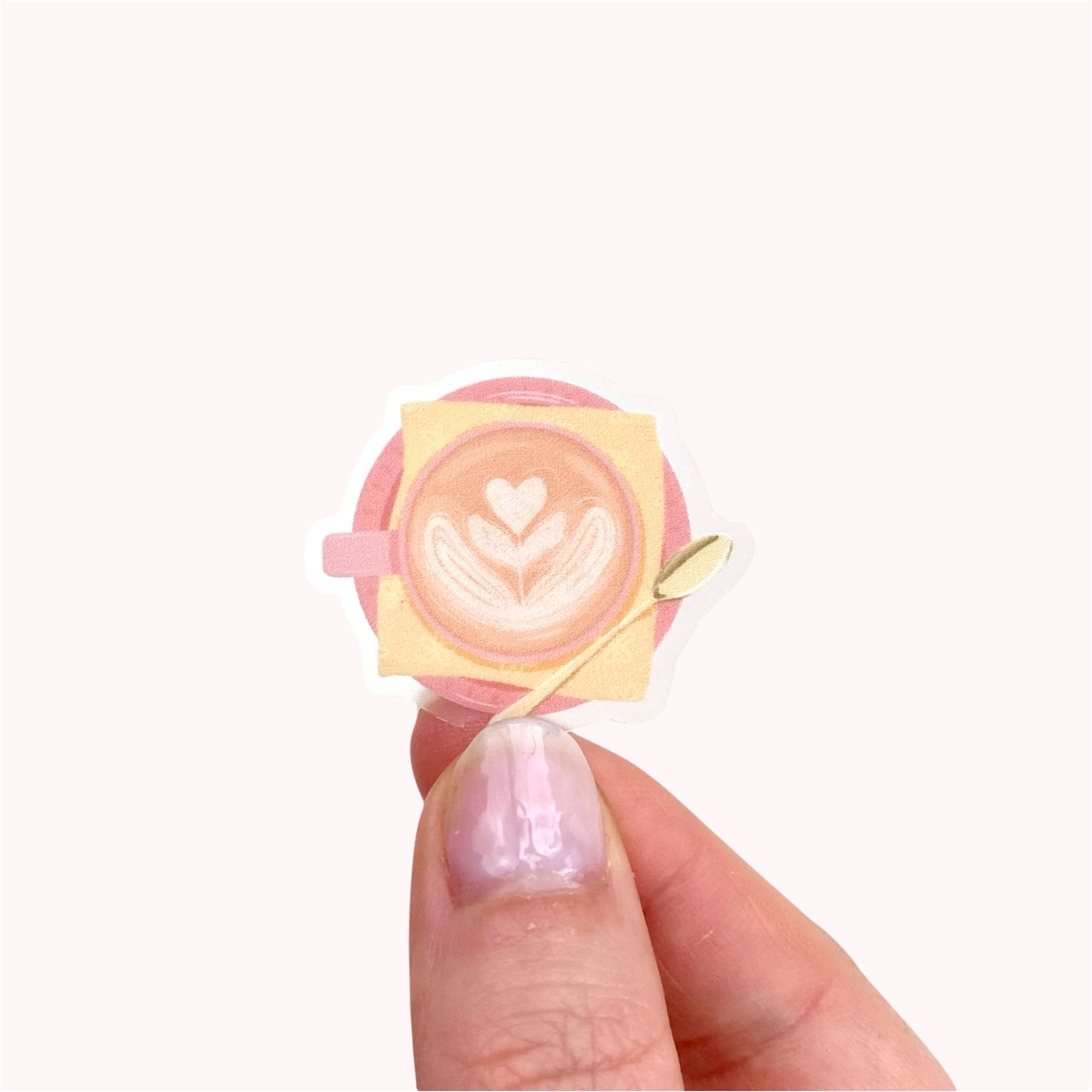 A transparent sticker with a detailed illustration of a latte from above with foamy heart shaped latte art, in a pink mug with a yellow napkin and gold teaspoon.