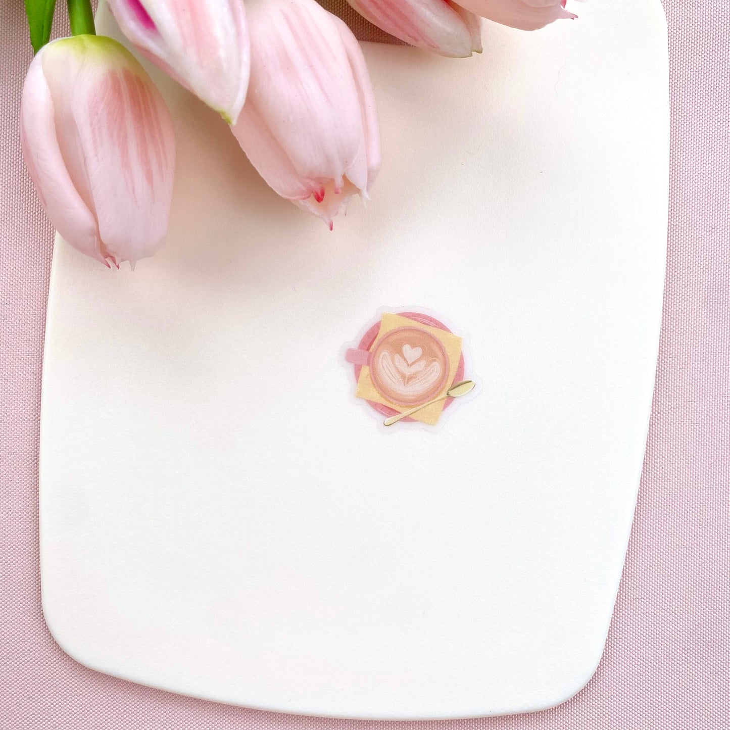 A transparent sticker with a detailed illustration of a latte from above with foamy heart shaped latte art, in a pink mug with a yellow napkin and gold teaspoon.