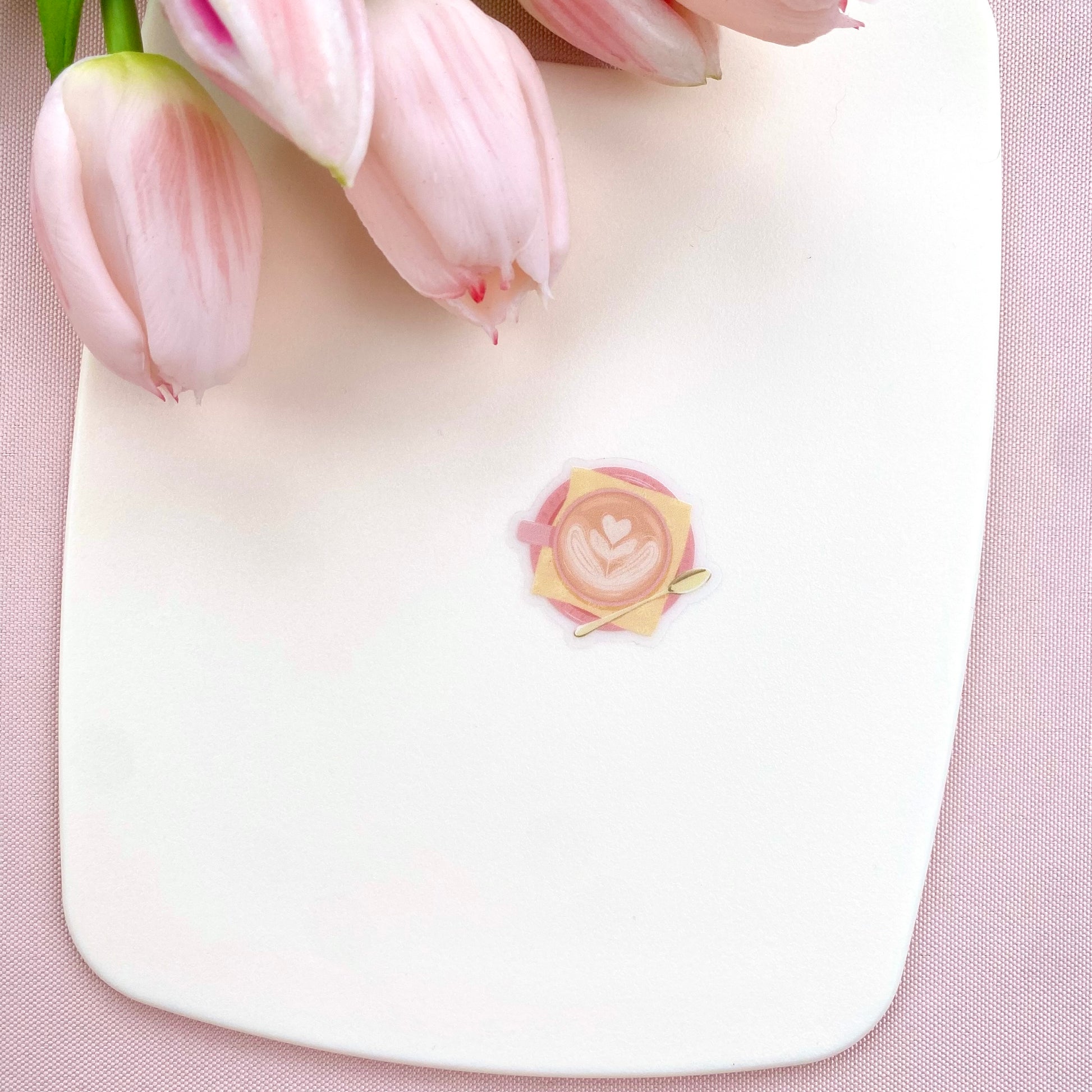 A transparent sticker with a detailed illustration of a latte from above with foamy heart shaped latte art, in a pink mug with a yellow napkin and gold teaspoon.