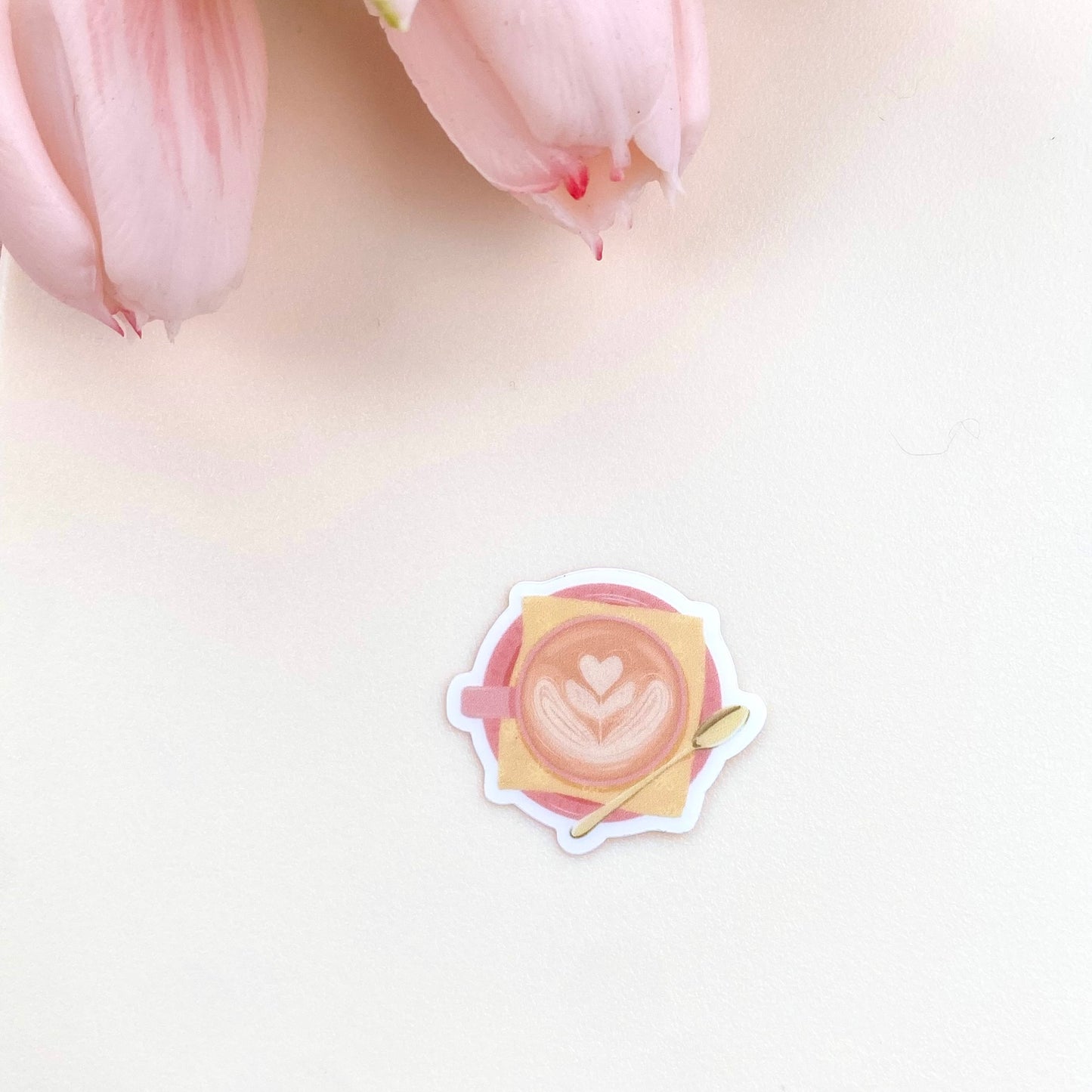 A transparent sticker with a detailed illustration of a latte from above with foamy heart shaped latte art, in a pink mug with a yellow napkin and gold teaspoon.