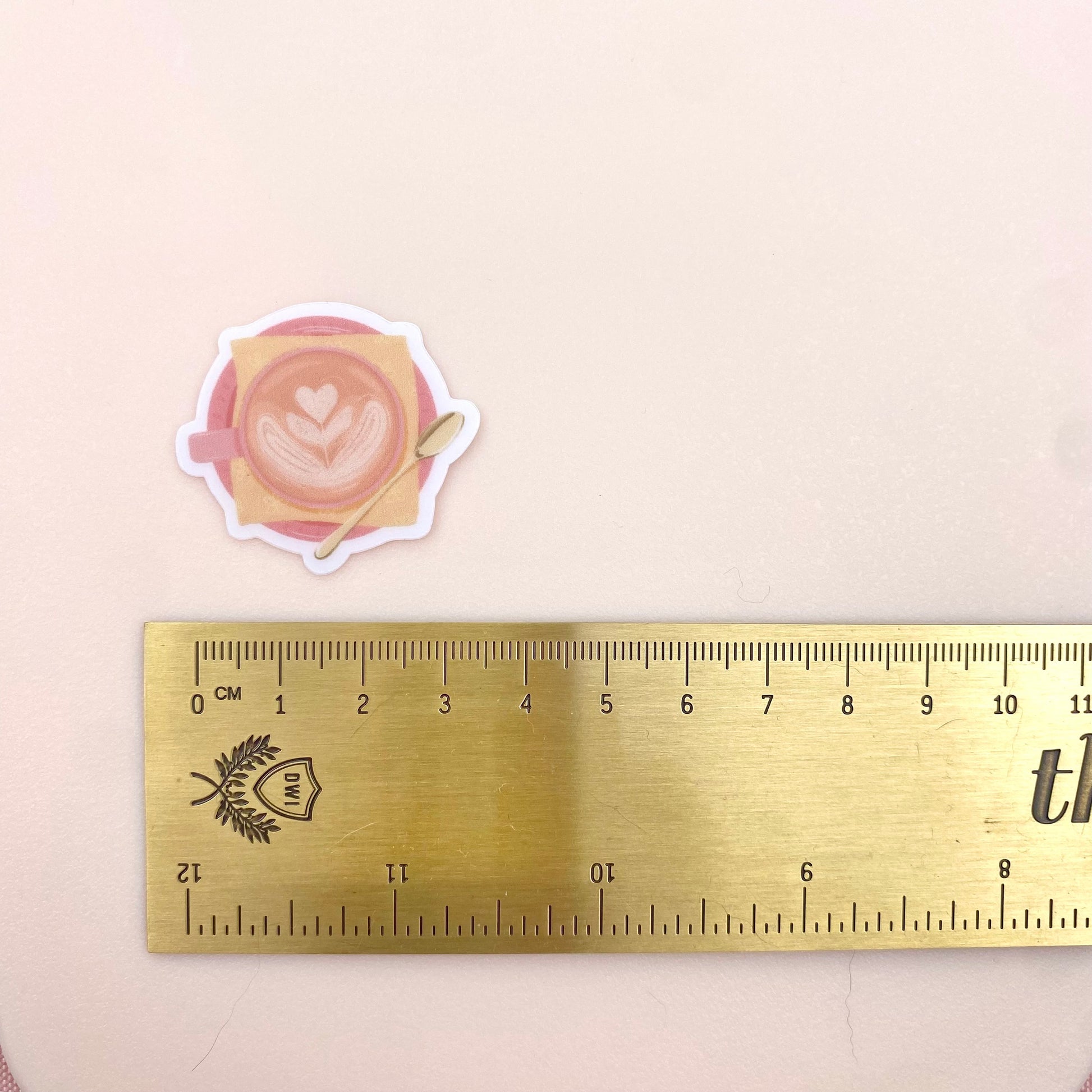 A transparent sticker with a detailed illustration of a latte from above with foamy heart shaped latte art, in a pink mug with a yellow napkin and gold teaspoon. Shown with a ruler for size reference.