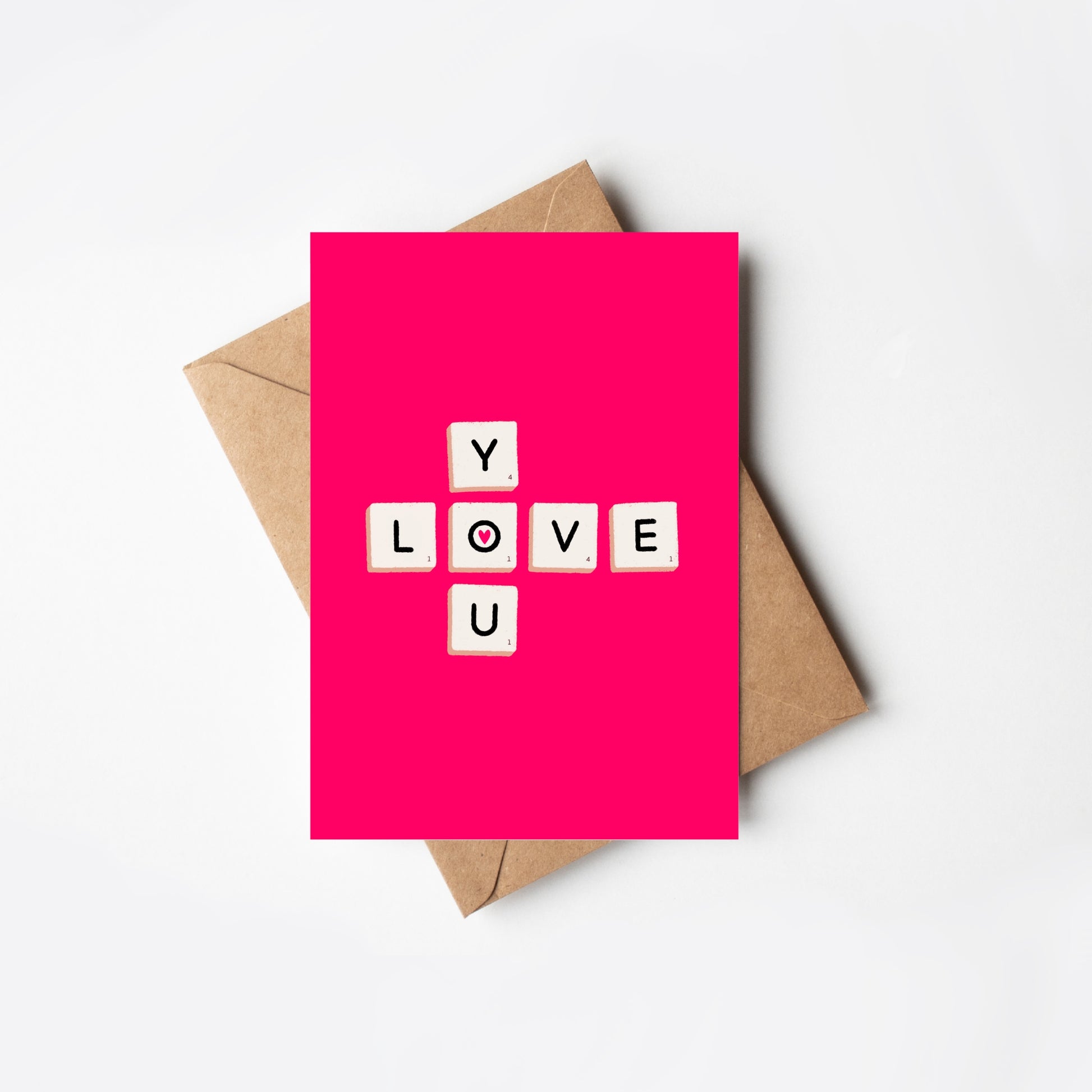 Pink card with 'LOVE YOU’ letter game tiles and a brown envelope on a white background