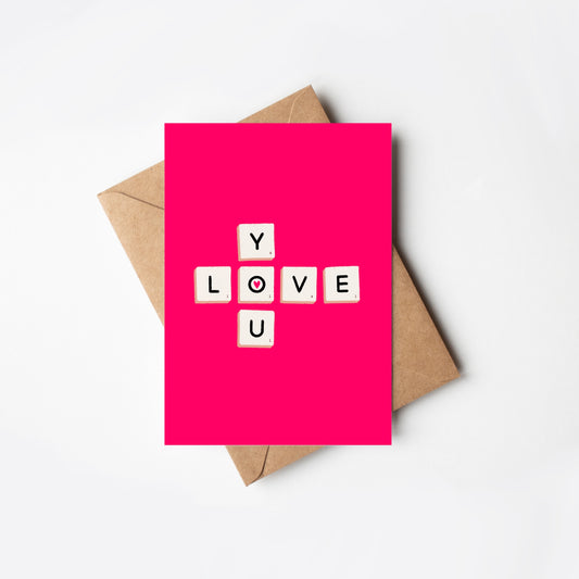 Pink card with 'LOVE YOU’ letter game tiles and a brown envelope on a white background
