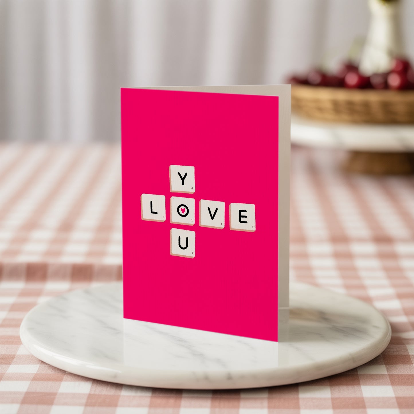 Pink card with 'LOVE YOU' in scrabble tiles on a marble stand with a checkered tablecloth background.